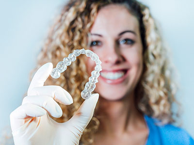The image shows a person holding up a clear dental retainer with their left hand while smiling at the camera.