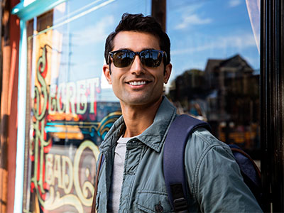 A young man wearing sunglasses, a backpack, and a jacket stands confidently in front of a storefront with a sign reading Sweet Treats.
